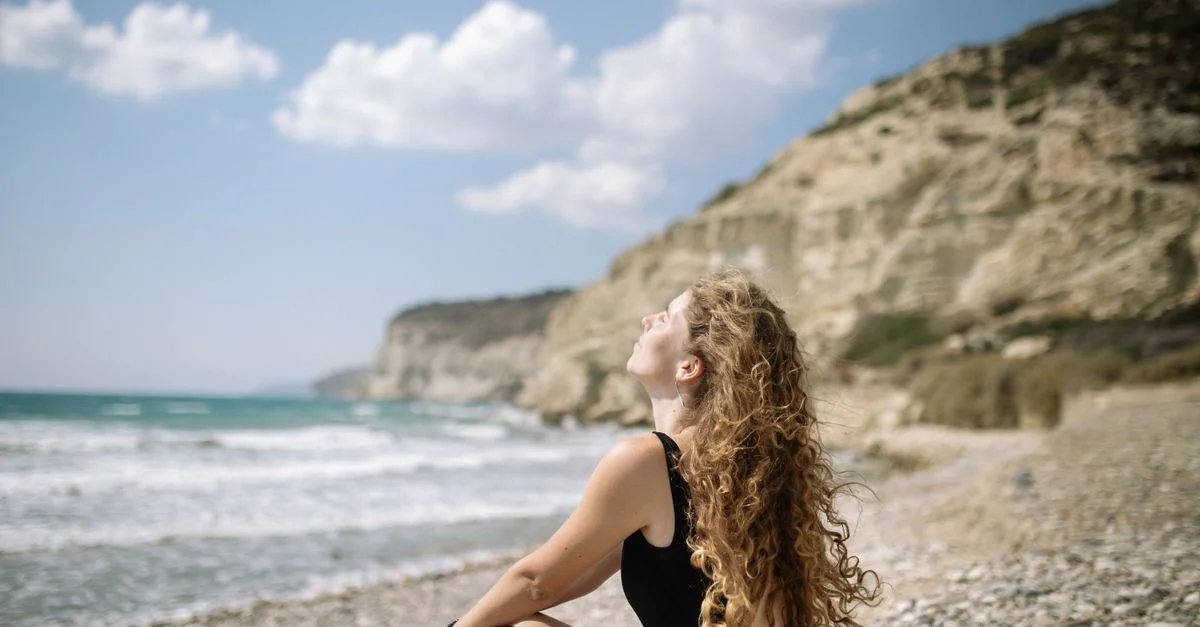 Una mujer con cabello rizado se sienta en una playa rocosa, disfrutando de un día soleado con vistas al océano.