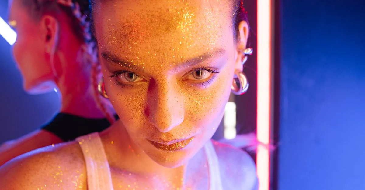 Retrato en primer plano de una mujer con maquillaje brillante, de pie bajo luces de neón vibrantes con pendientes visibles.