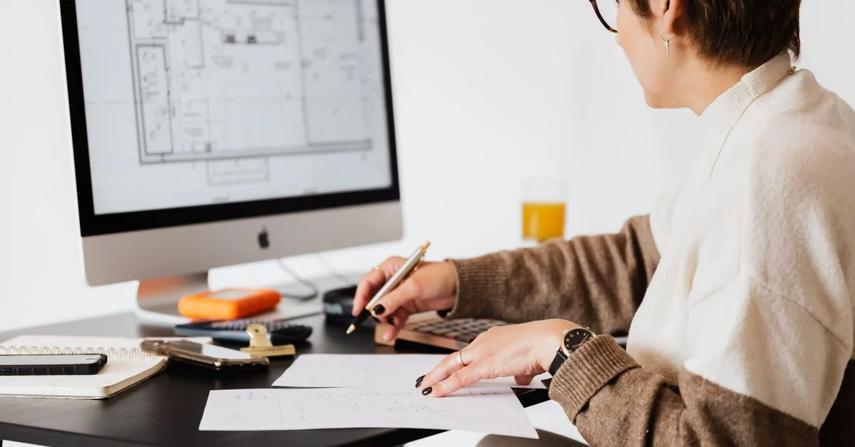 Mujer diseñando planos arquitectónicos con ordenador de escritorio en un entorno de oficina moderna.
