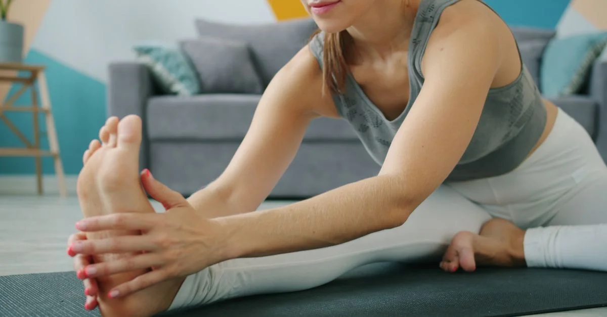 Una mujer realizando una postura de yoga de flexión hacia adelante sentada sobre una esterilla en el interior de una vivienda.
